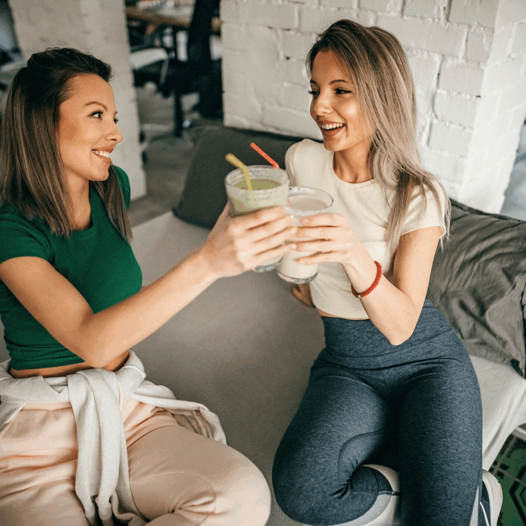 Dvije žene u sportskoj odjeći sjede na kauču i nazdravljaju čašama s Herbalife shakeovima nakon treninga, nasmijane i pune energije.