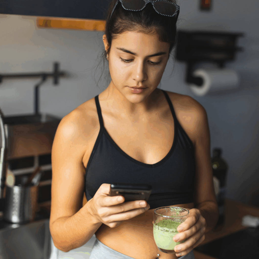 Mlada žena u sportskoj odjeći stoji u kuhinji i drži čašu s Herbalife shakeom dok gleda u mobitel, planirajući svoj zdravi doručak i fitness rutinu.