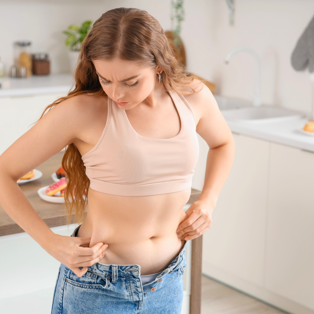 Žena u sportskoj odjeći stoji u kuhinji i promatra trbuh, provjeravajući višak masnih naslaga tijekom procesa mršavljenja.