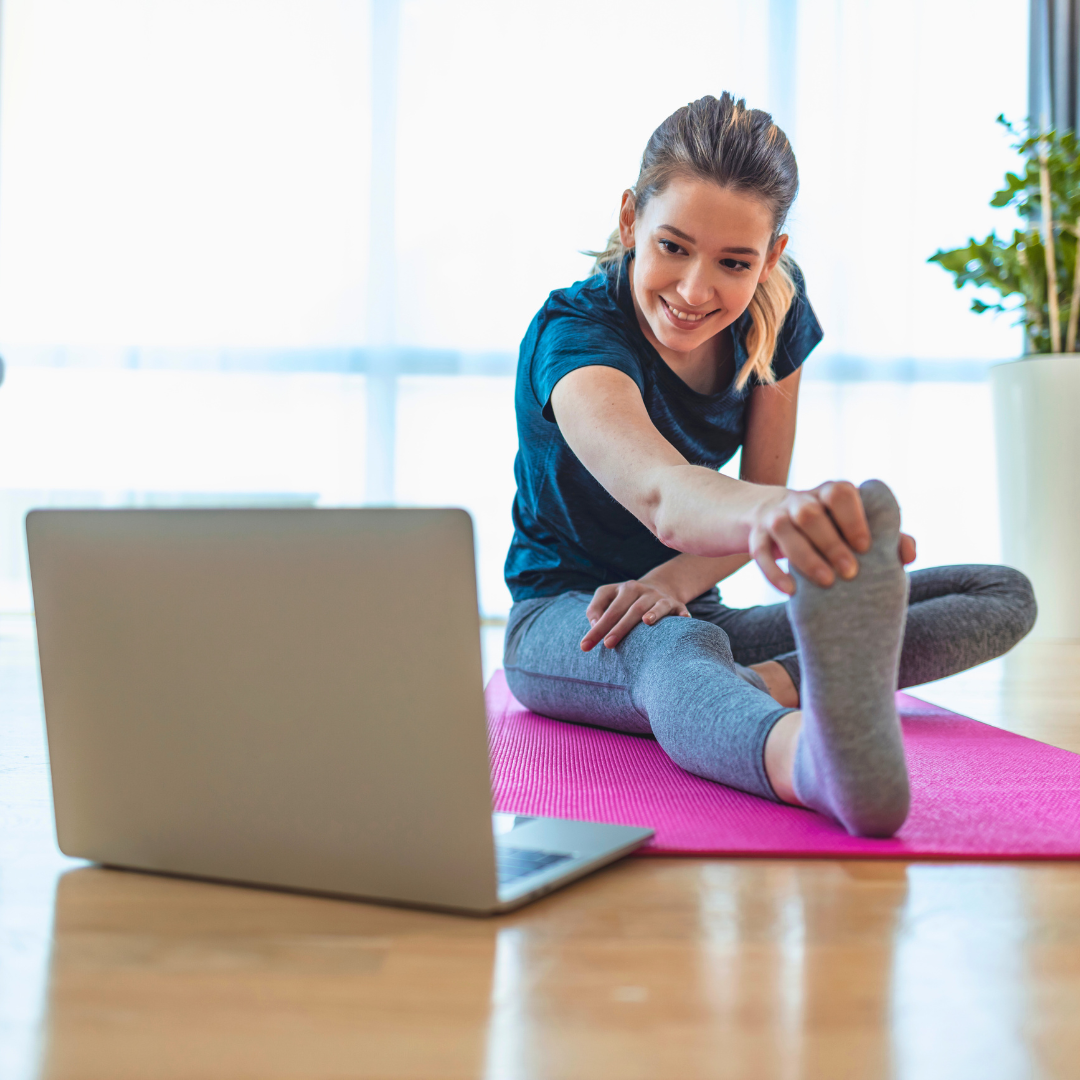 Žena se smije dok radi vježbe istezanja na ružičastoj prostirci ispred laptopa, prateći online trening od kuće.