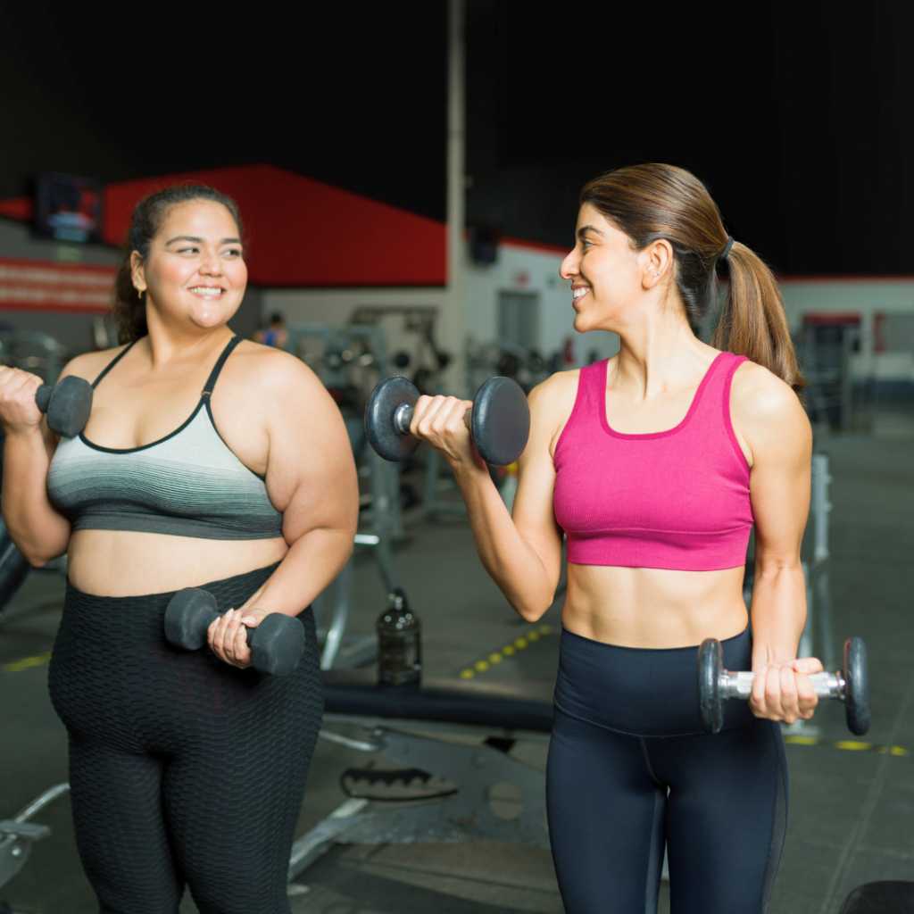 Dvije žene vježbaju zajedno u teretani i nasmijano podižu utege, prikazujući podršku, zajedništvo i motivaciju tijekom treninga.