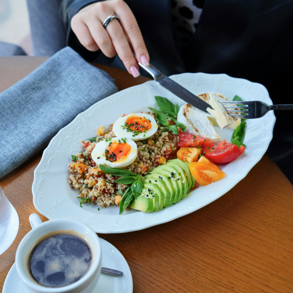 Zdravi doručak na tanjuru s avokadom, kuhanim jajima, kvinojom, rajčicama i sirom, uz šalicu kave, prikazuje uravnotežen i nutritivno bogat obrok.