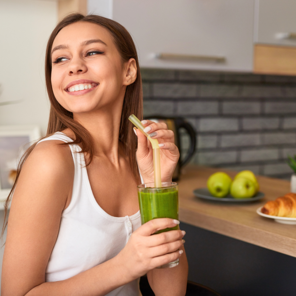 Nasmijana žena u bijeloj majici drži čašu zelenog smoothieja u kuhinji, simbolizirajući zdrav početak dana i svjesne prehrambene navike.