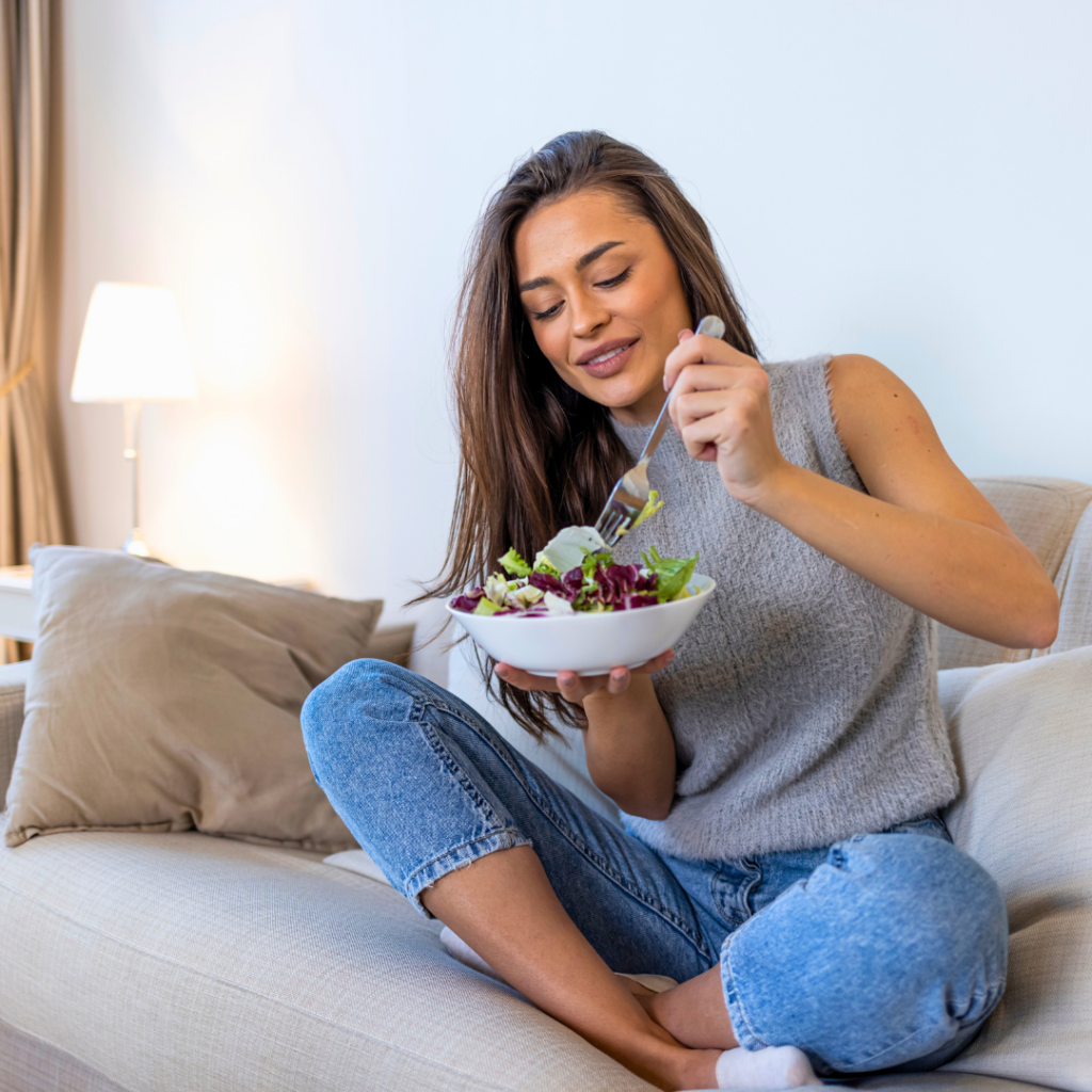 Žena sjedi opušteno na kauču i jede svježu zelenu salatu iz zdjele, prikazujući uravnotežen i zdrav način života.