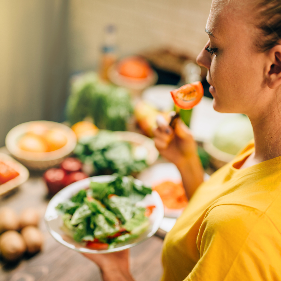 Žena u žutoj majici jede svježu salatu i krišku rajčice u kuhinji punoj povrća, prikazujući naviku zdrave i uravnotežene prehrane.