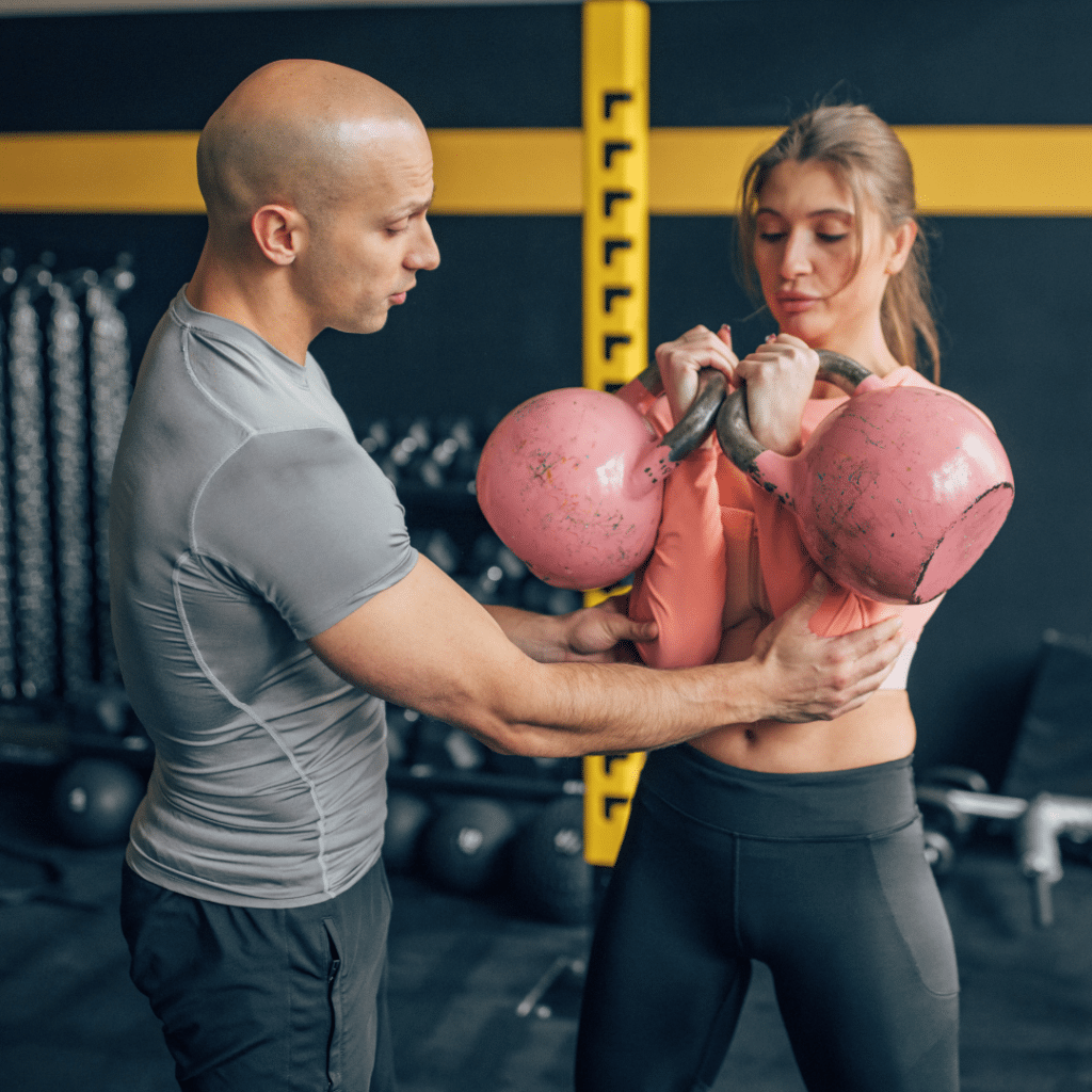 Trener u teretani pomaže ženi tijekom vježbe s dvije girje, ispravljajući tehniku i usmjeravajući pokret za pravilno izvođenje.