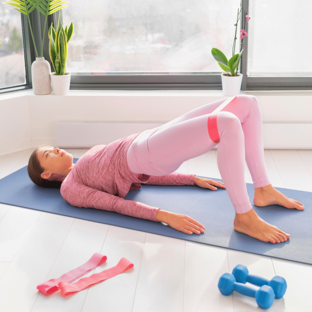 Žena u ružičastoj sportskoj odjeći izvodi most na prostirci koristeći elastičnu traku za otpor, fokusirana na jačanje mišića stražnjice i nogu kod kućnog treninga.