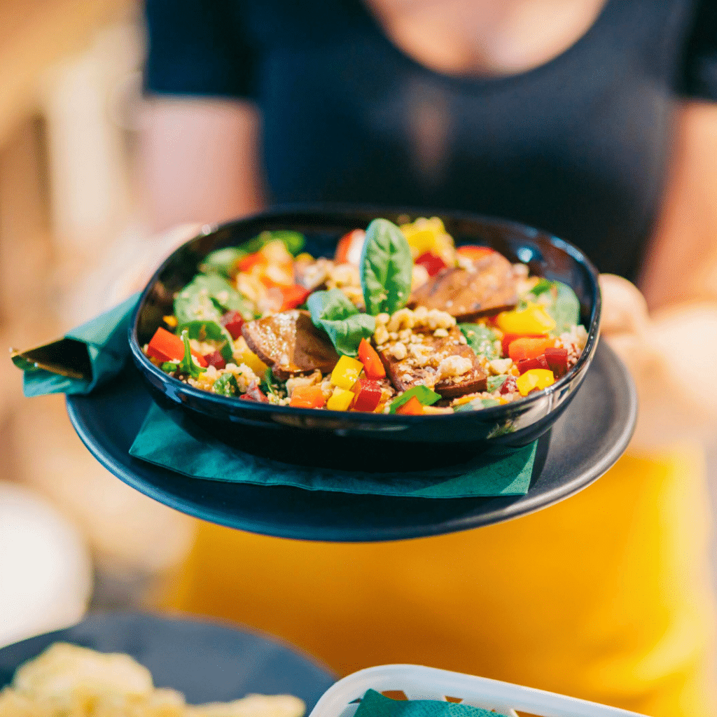 Poslužena veganska zdjela s komadićima tofua, paprikom u boji, špinatom i orašastim posipom, aranžirana u crnoj zdjeli na tanjuru s tamnozelenim ubrusom.
