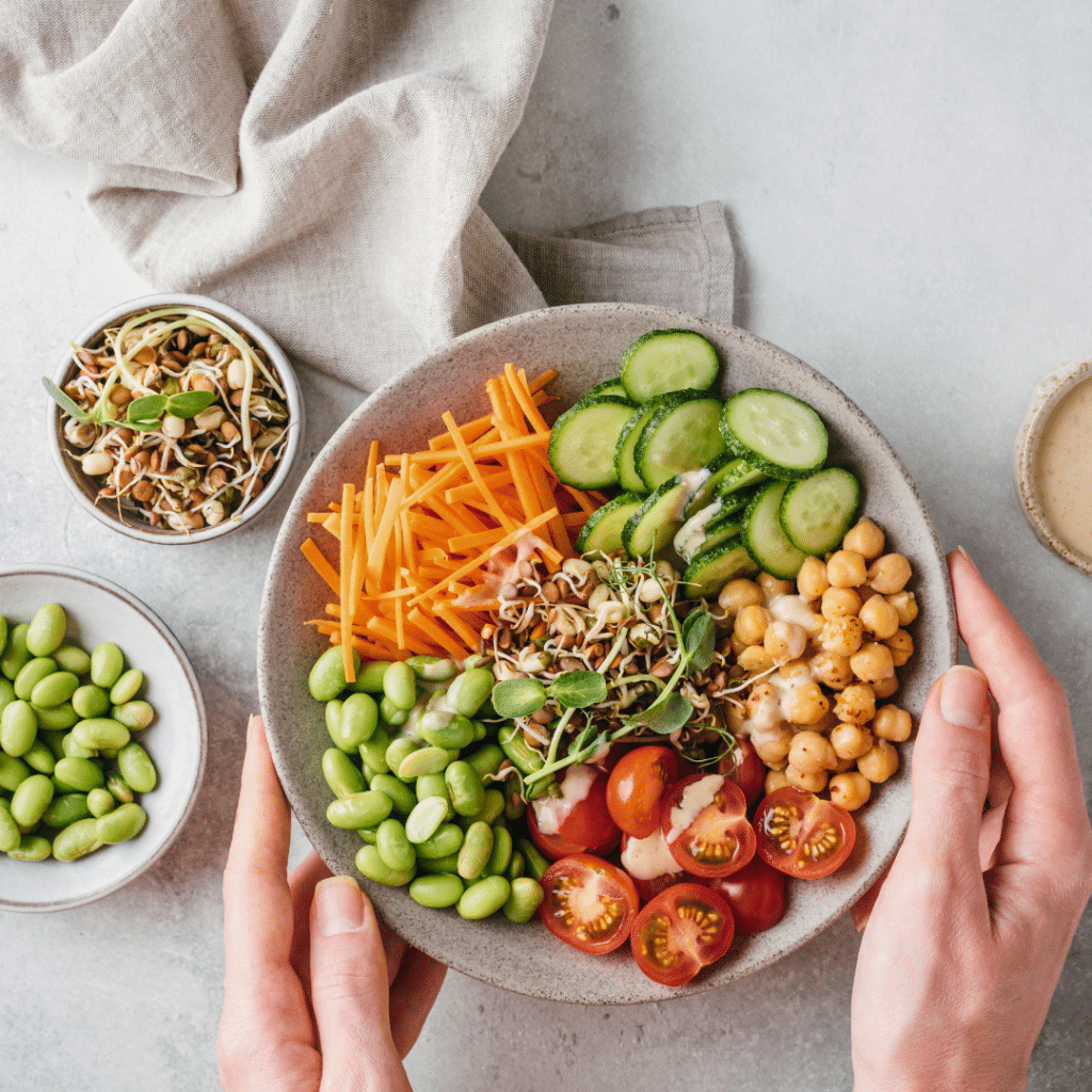 Veganska zdjela s raznobojnim povrćem — slanutak, rajčice, edamame, krastavci, naribana mrkva i klice, preliveno laganim umakom i posluženo u keramičkoj zdjeli.