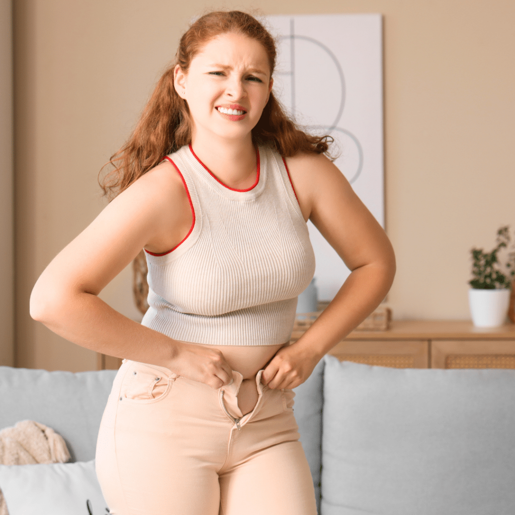 Žena se muči zakopčati hlače – prikaz frustracije zbog viška kilograma prije uspješne transformacije uz BudiFit program mršavljenja.