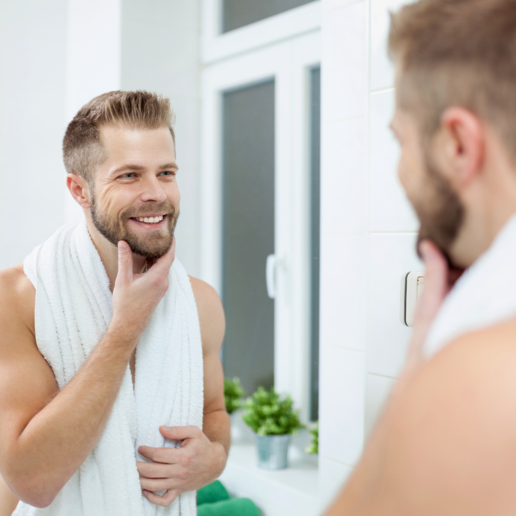 Muškarac nakon njege lica promatra svoj odraz u ogledalu i zadovoljno se smije – simbol zdravije, čvršće i hidratizirane kože zahvaljujući vitaminu C i bakuchiolu.