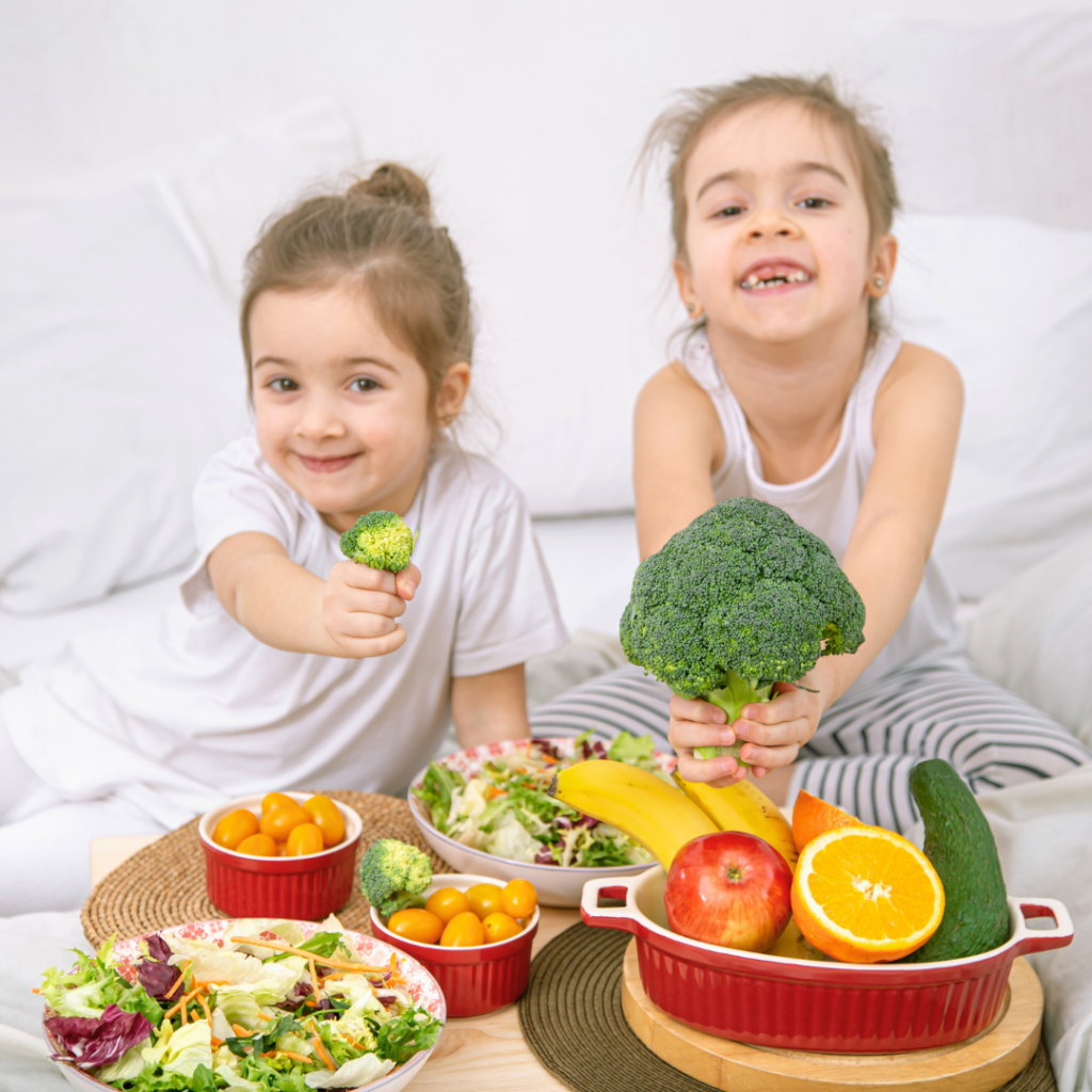 Djeca s osmijehom drže brokulu i svježe voće – primjer zdrave užine bogate povrćem i voćem koja potiče pravilne prehrambene navike kod djece.
