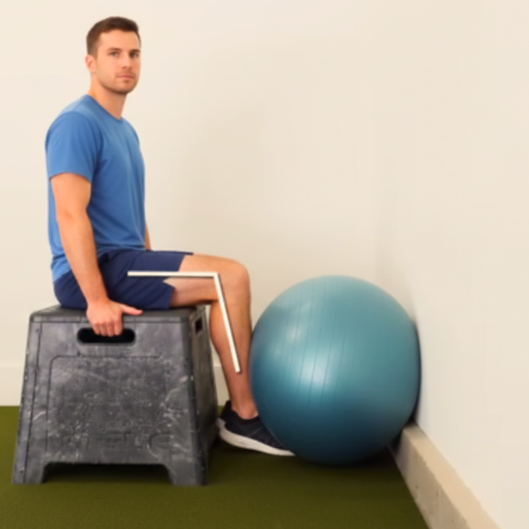 Izometrička ekstenzija koljena nakon ACL operacije – muškarac sjedi na povišenju i izvodi statičko opružanje koljena pod kutom od 90 stupnjeva uz terapijsku loptu, vježba za jačanje kvadricepsa u ranoj fazi rehabilitacije.